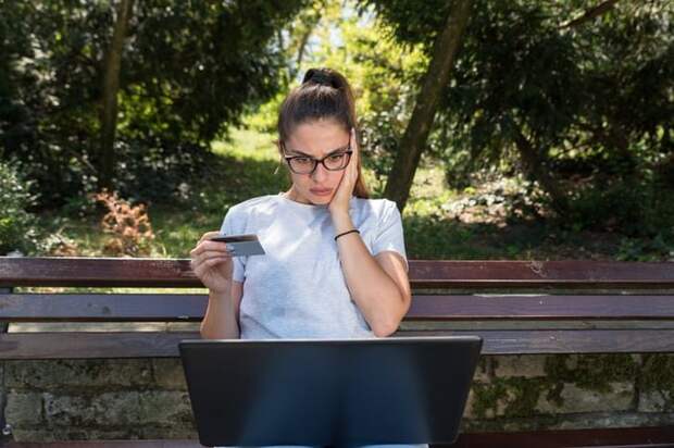 A young woman holding a credit card looks surprised while she checks a laptop.