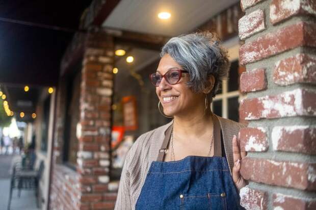 A smiling person in an apron outside a building.