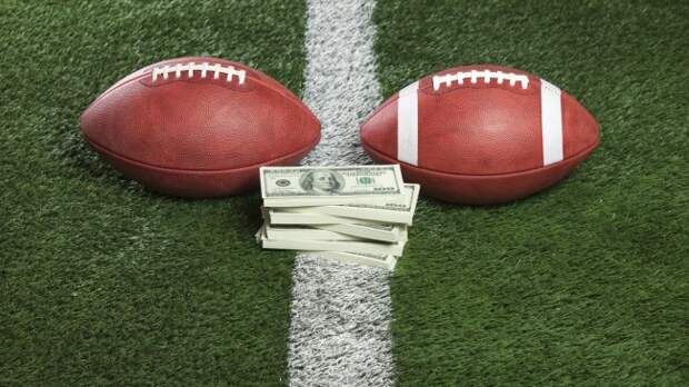 An NFL and NCAA football rest next to a stack of cash on the field.