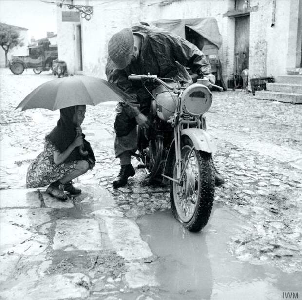 Маленькая девочка с зонтиком наблюдает за попыткой британского гонщика отремонтировать карбюратор мотоцикла под проливным дождем. Италия. 4 октября 1943 года
