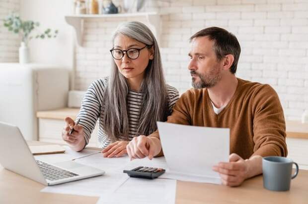 A mature couple sitting at a kitchen table with papers, a laptop, and a calculator.