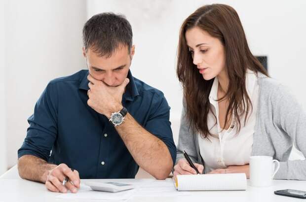 Man holding pen and looking in calculator next to woman writing in notebook