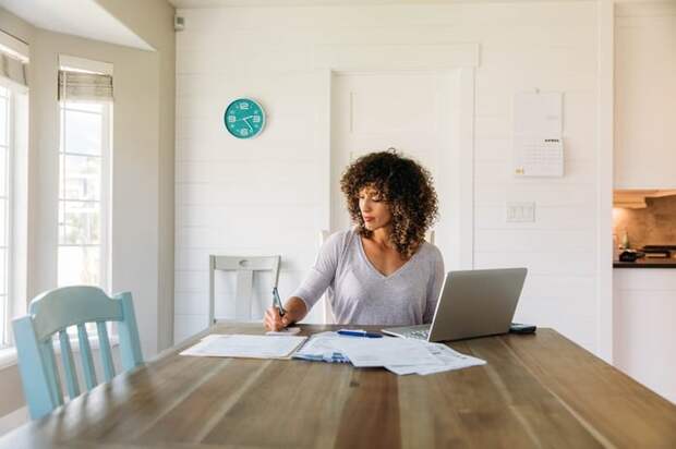 A woman sitting at a sunny kitchen table with her laptop and notes in front of her.