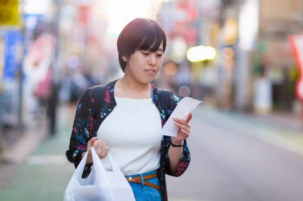 A person walking down the street with a grocery bag while reading a receipt.