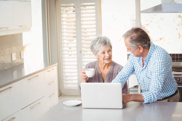Senior couple at laptop in kitchen; woman is holding a mug and man is typing