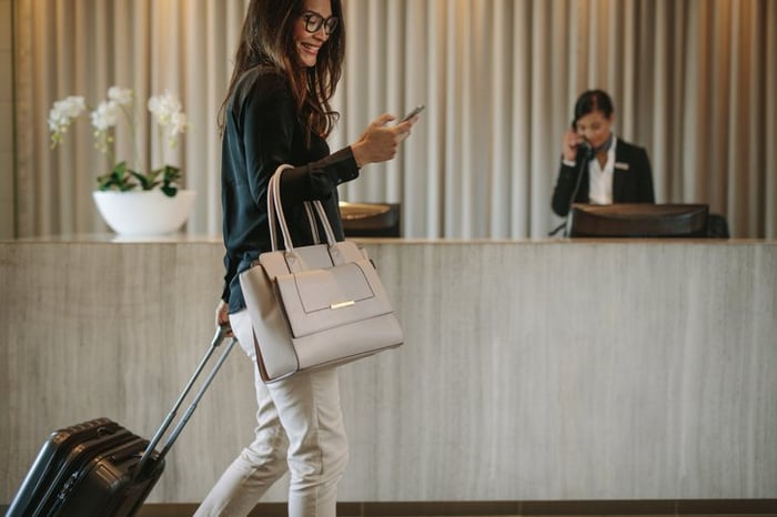 A smiling woman talking on her phone while walking with her suitcase into the lobby of a hotel.