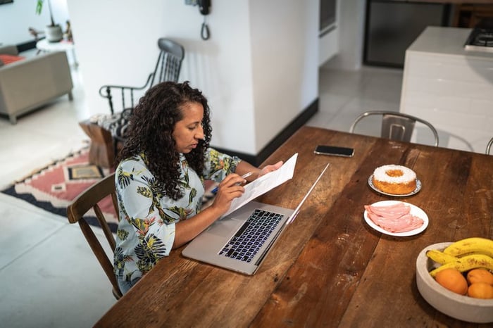 A person manages their home finances using documents and a laptop at their kitchen table.