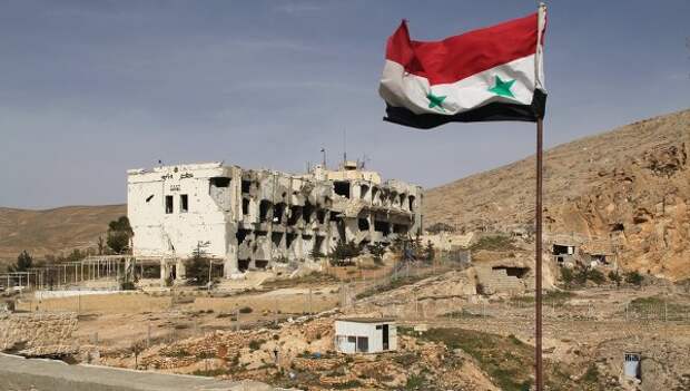 Сирийский флаг на фоне разрушенного дома в сирийском городе Маалюля в 55 км от Дамаска, который дважды захватывали и грабили боевики из группировки Джабхат ан-Нусра