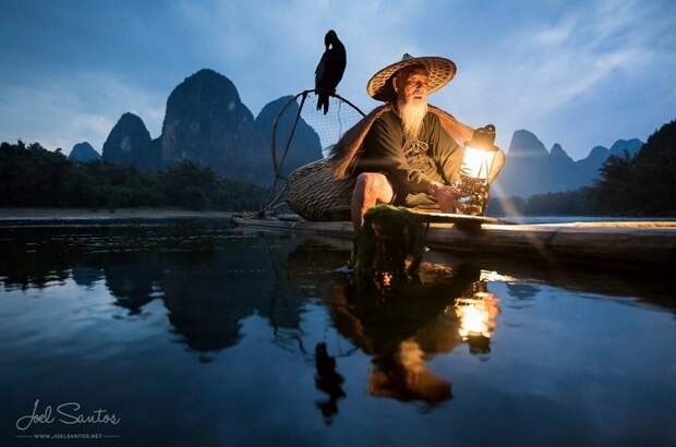 Китайский рыбак, на рыбалке на горном озере, в узком бревенчатом плоту, с корзиной для рыбного улова и сидящим бакланом.