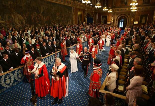 Под звуки фанфар королева с супругом входят в здание парламента. Их сопровождают официальные лица, пажи и герольдмейстеры - специальные распорядители королевских церемоний. На фото: Елизавета II с супругом принцем Филипом направляются в зал заседаний палаты лордов, 2014 год