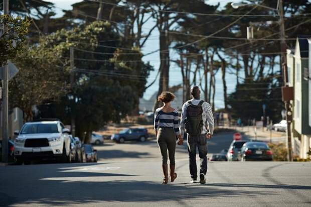 Two people holding hands while walking down the middle of a street surrounded by trees and parked cars.