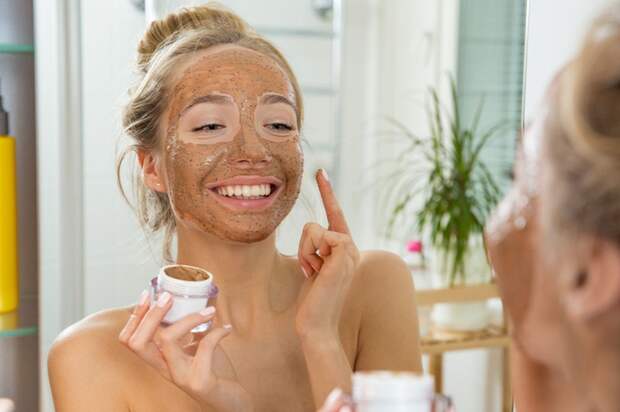 Young beautiful girl applying facial scrub mask on skin. Looking in the mirror in bathroom, Wrapped in a towel, having fun.