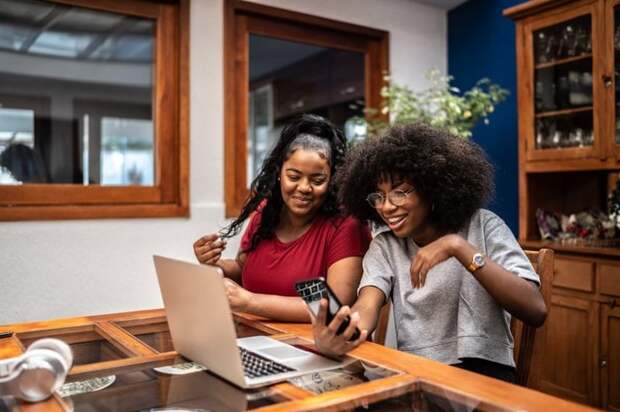 Two smiling people sitting in their kitchen and looking at a laptop while one holds a phone.