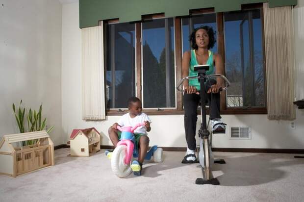 A parent uses an exercise bike at home while their young child sits on their own tricycle.