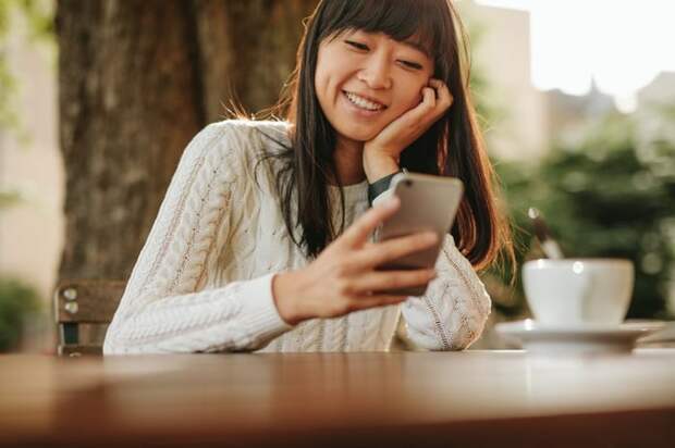 A smiling person sitting at an outdoor cafe table and looking at their cellphone.