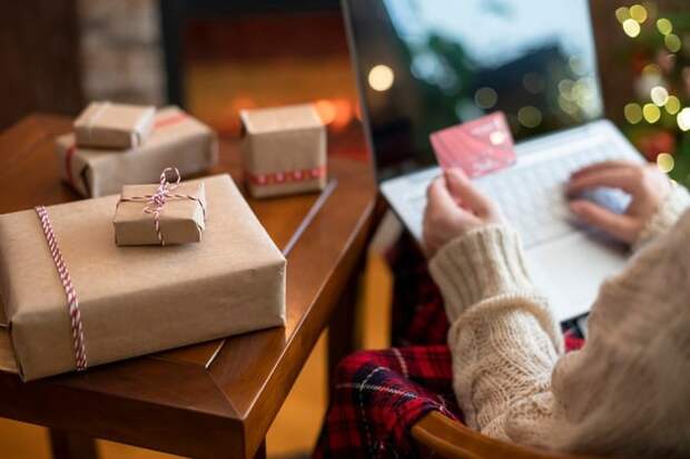 A person holding a credit card and online shopping on their laptop from their couch with wrapped gifts next to them.
