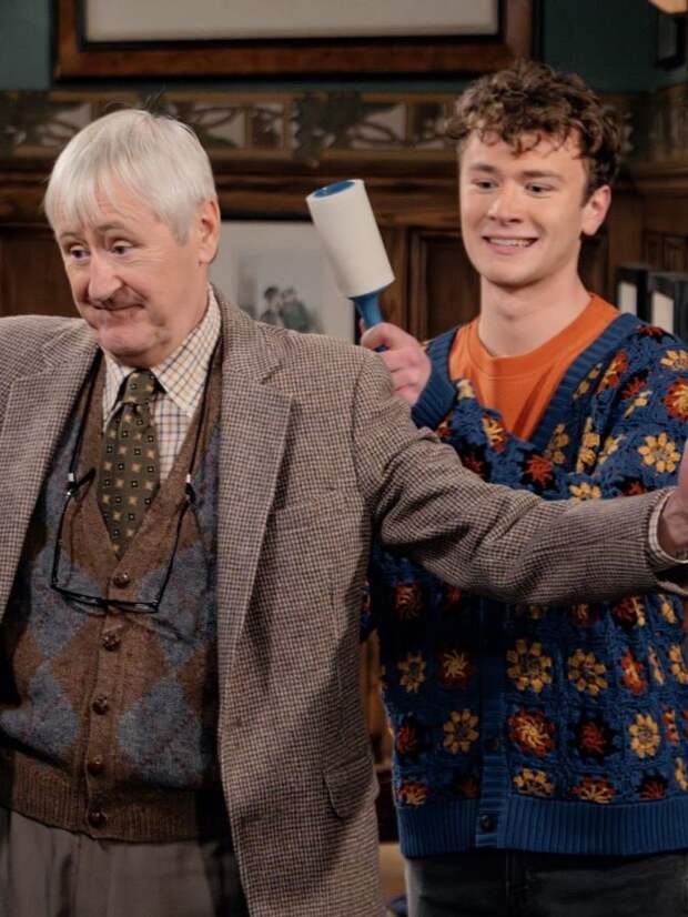 Alan holds his arms out while David uses a lint brush on them