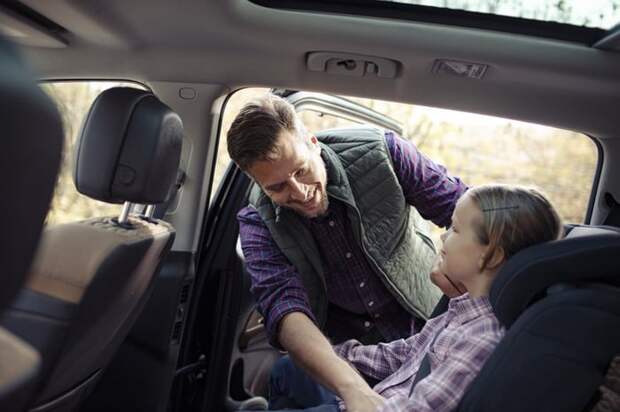 A smiling father helping his young daughter into a carseat.